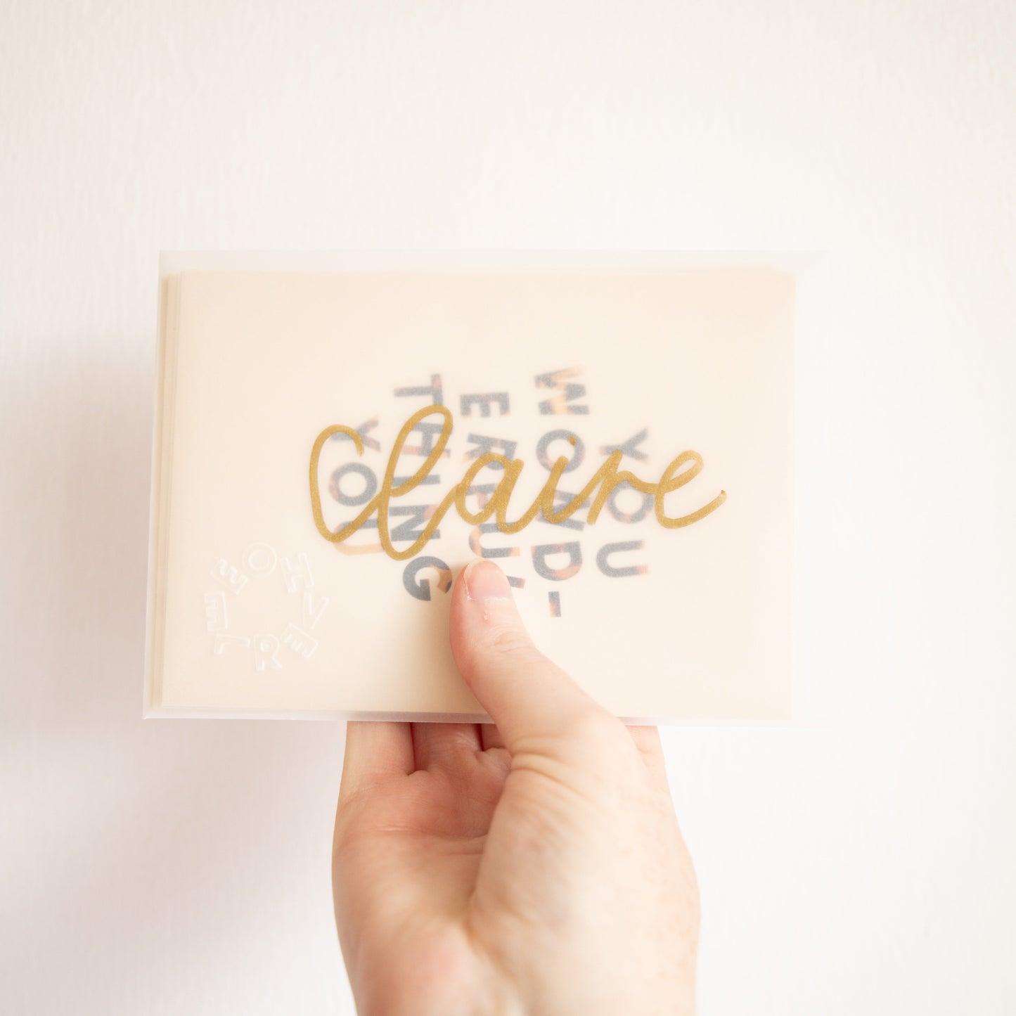 Hand holding an A6 postcard in a transparent envelope with gold hand-lettered text in front of a white wall