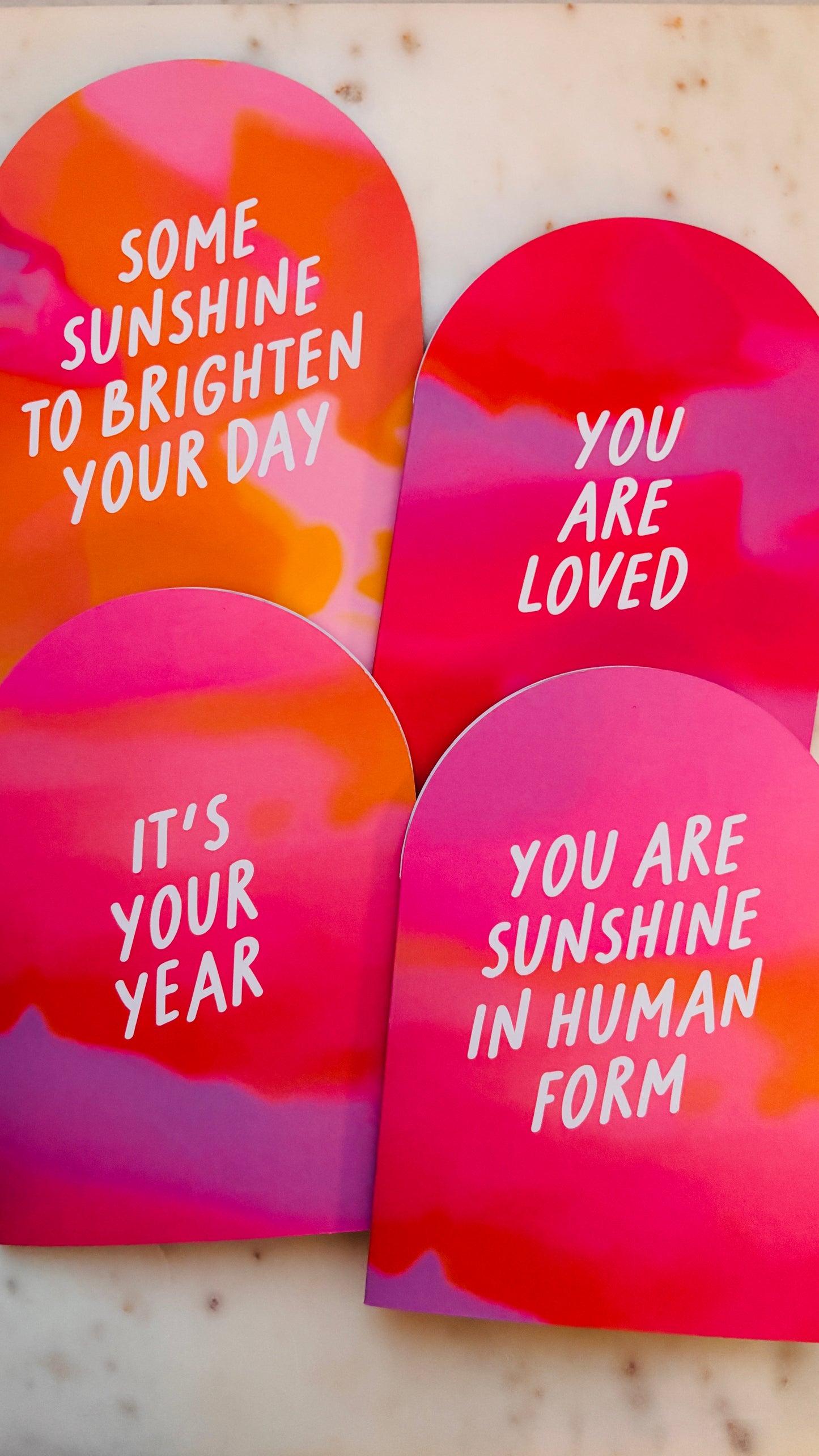 Four colourful A6 Arch Cards with pink and orange gradient backgrounds. The wording on the cards is: 'Some sunshine to brighten your day', 'you are loved', 'it's your year' and 'you are sunshine in human form'. The cards are on a marble surface.