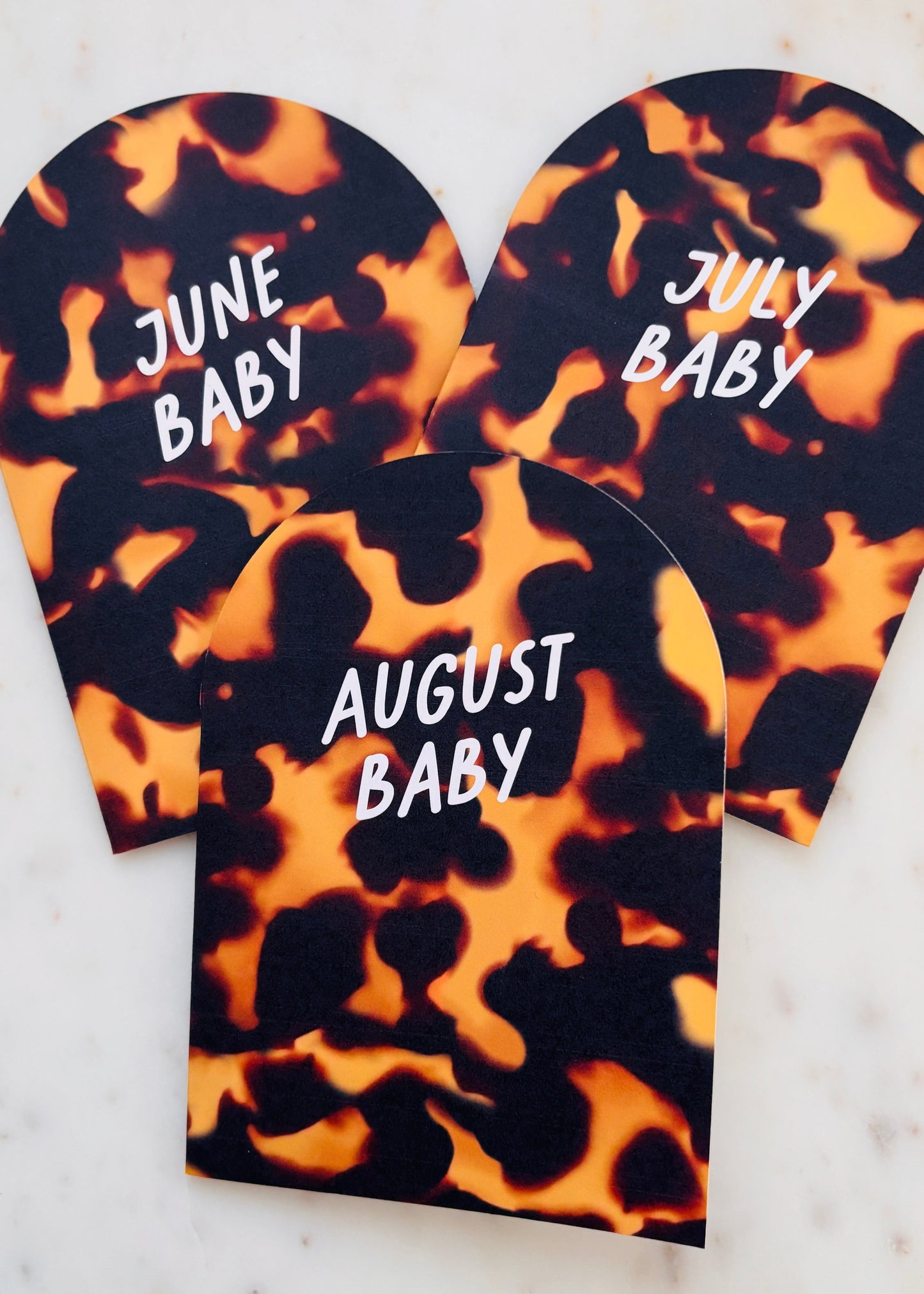 Three tortoiseshell-patterned A6 Arch Cards with white  'June Baby', 'July Baby', and 'August Baby' on each of them. The cards are on a marble background.