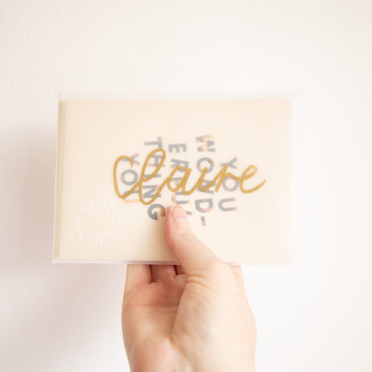 Hand holding an A6 postcard in a transparent envelope with gold hand-lettered text in front of a white wall