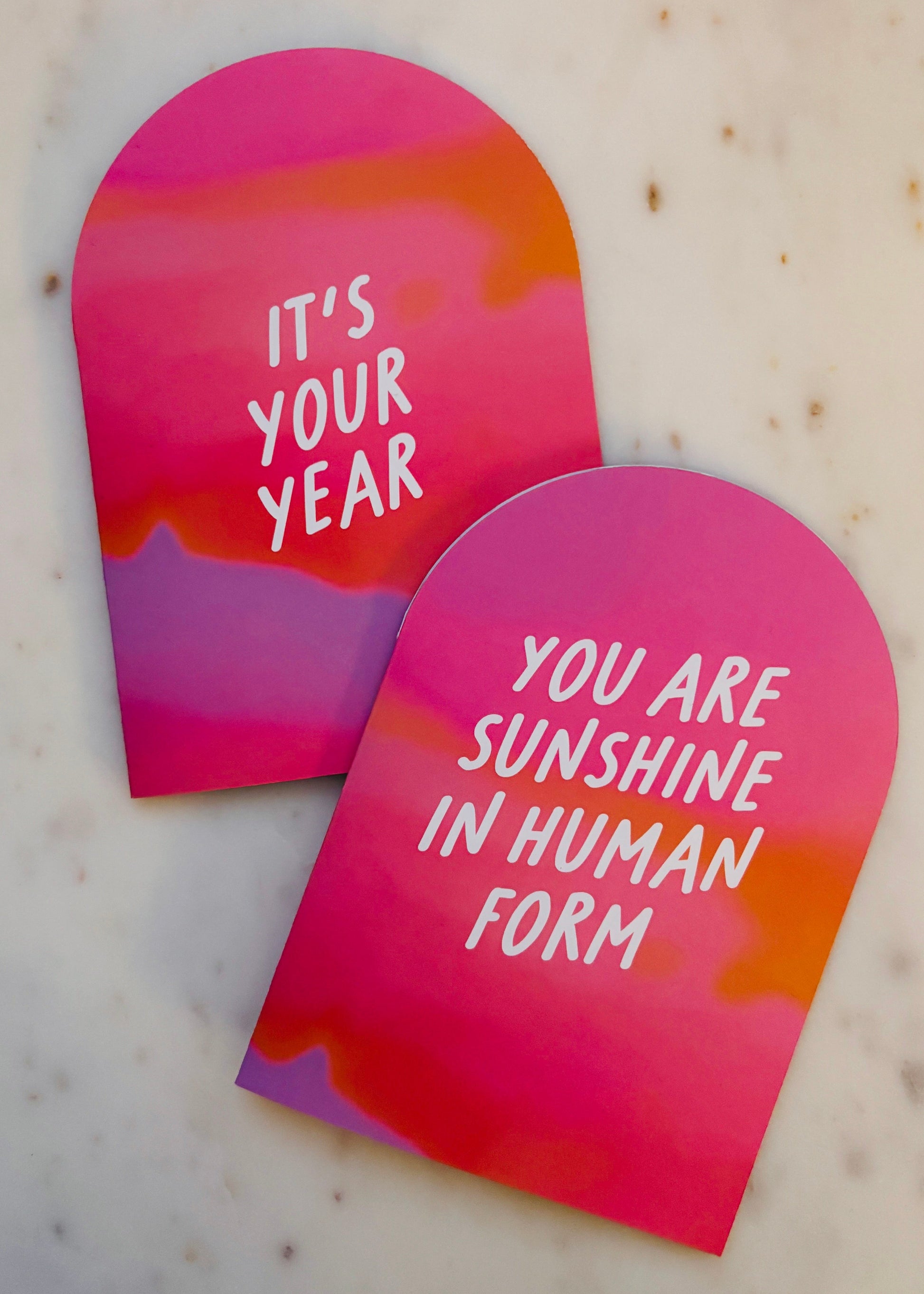 Two A6 Arch cut pink and orange gradient greetings cards with motivational messages on a marble surface.