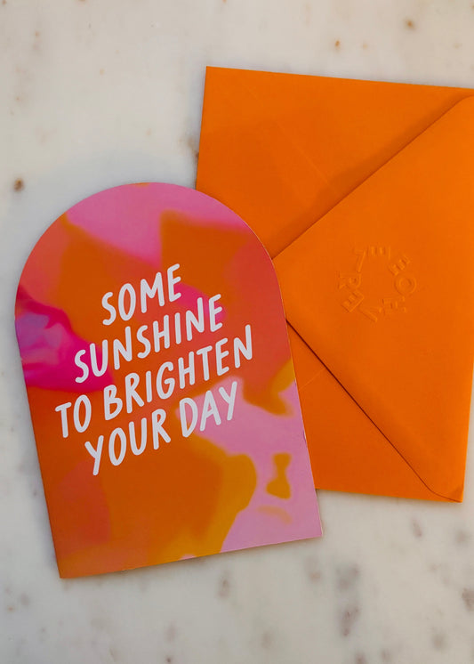 A6 Arched orange and pink gradient greetings card with 'Some sunshine to brighten your day' text and an orange envelope on a marble surface.