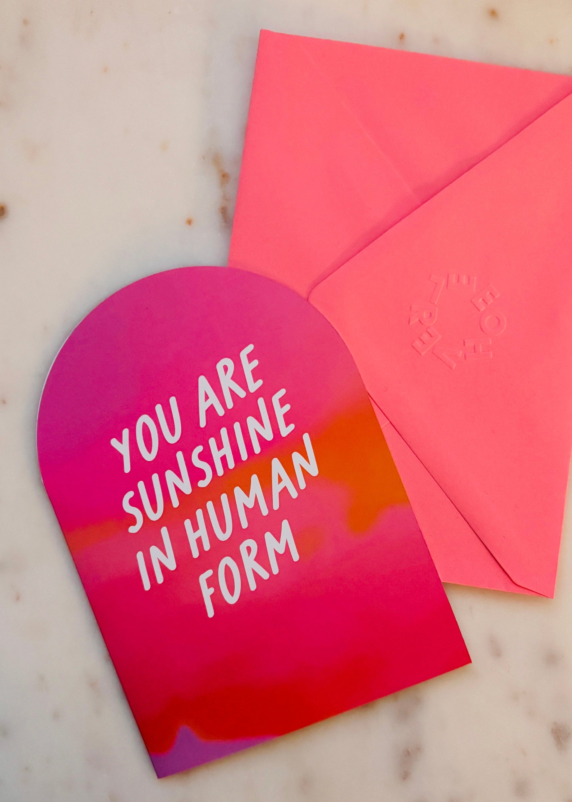 An arch A6 greetings card with 'You are sunshine in human form' text on a pink and orange gradient, with a pink envelope. On a marble background.